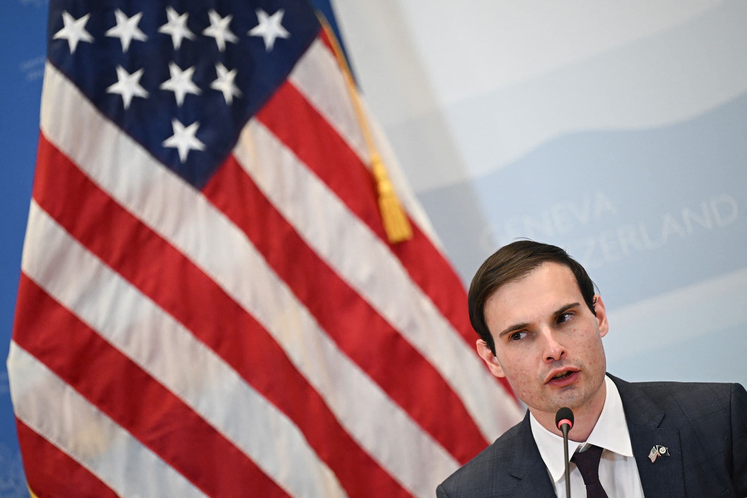 A man in a suit speaking into a microphone in front of an American flag.